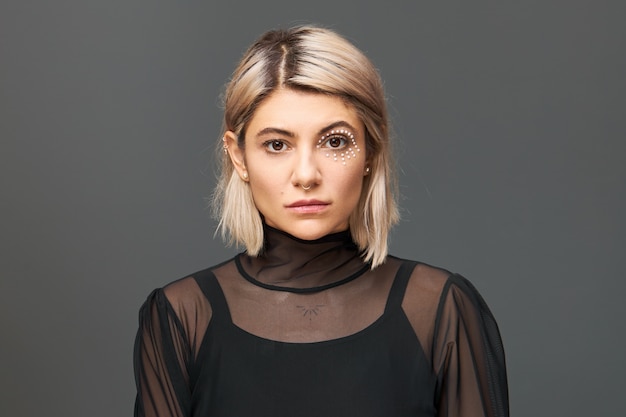 Horizontal image of amazing young female with blonde bob hairstyle and artistic make up with white crystals around her eye posing isolated in trendy blouse, having confident look