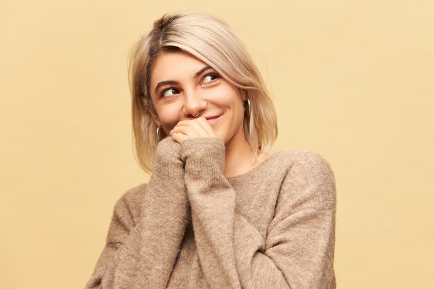 Horizontal image of attractive charming young Caucasian female wearing nose ring, round earrings and stylish warm beige sweater holding hands together and looking away with mysterious smile