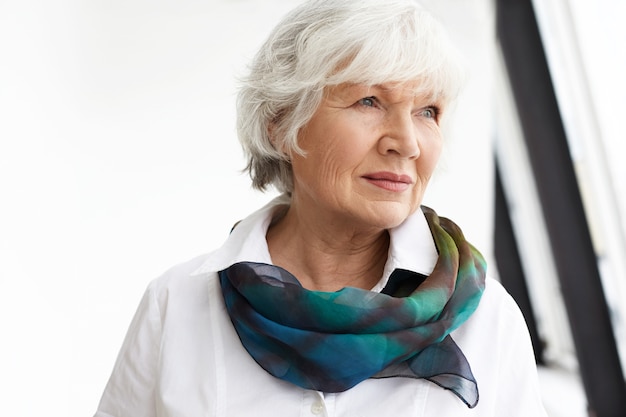 Horizontal image of attractive fashionable retired female wearing white shirt and elegant silk scarf daydreaming at home,