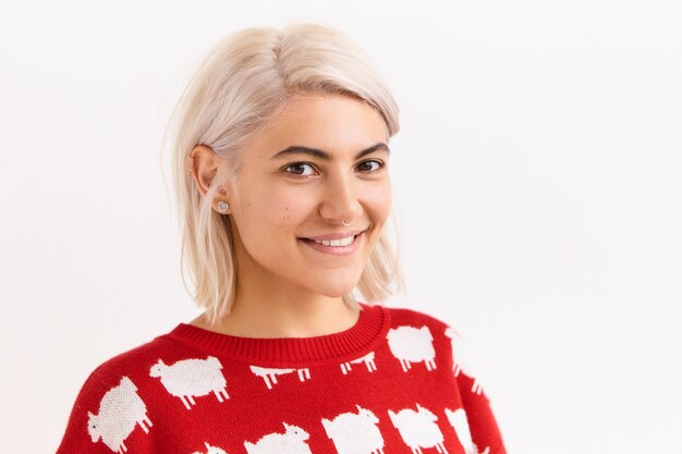 Horizontal image of beautiful European student girl rejoicing at good positive news, gets A mark, posing isolated in red sweater, with cute adorable smile, showing straight teeth