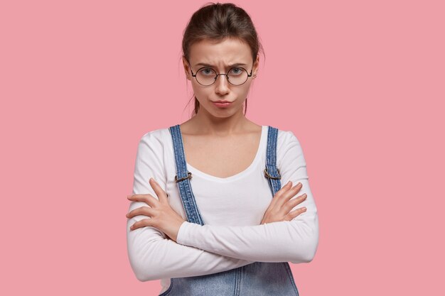 Horizontal image of displeased dejected young woman looks with sullen expression, crosses hands over chest, being in bad mood