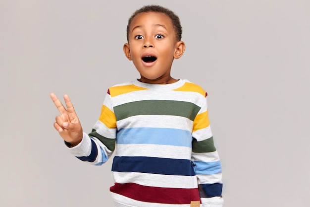 Horizontal image of funny excited African boy keeping mouth wide opened being surprised to see something unexpected, making peace gesture. Emotional black child showing victory sign and exclaiming