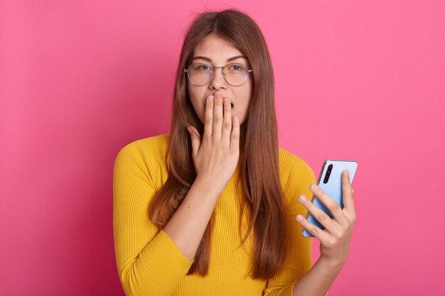 Horizontal image of impressed emotional cute young female wearing eyeglasses and yellow sweatshirt, covering mouth with hand, holding smartphone, looking directly at camera. Technology concept.