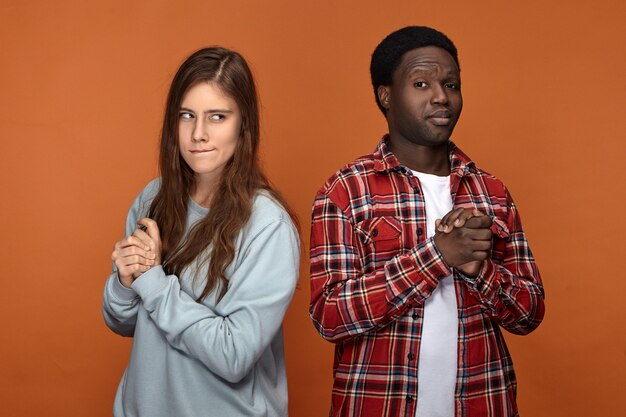 Horizontal image of interracial couple pulling tricks on each other, posing isolated against blank orange  wall , looking suspicious, planning trick or prank, rubbing hands