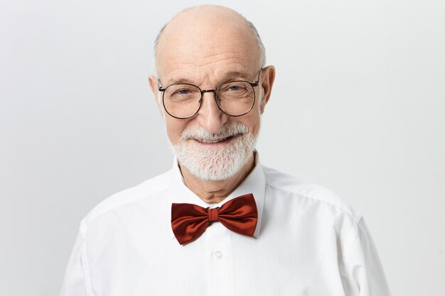 Horizontal image of successful handsome bald bearded elderly male dressed in stylish clothes posing at blank wall, rejoicing at good news, looking  with beaming confident smile