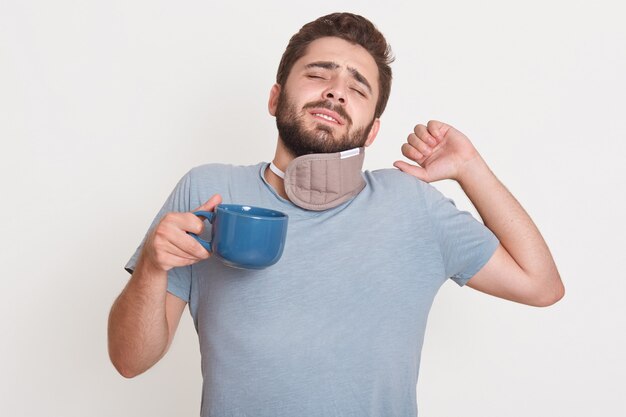 Horizontal image of tired bearded man standing isolated over white wall, closing eyes, raising one hand, having bad sleep