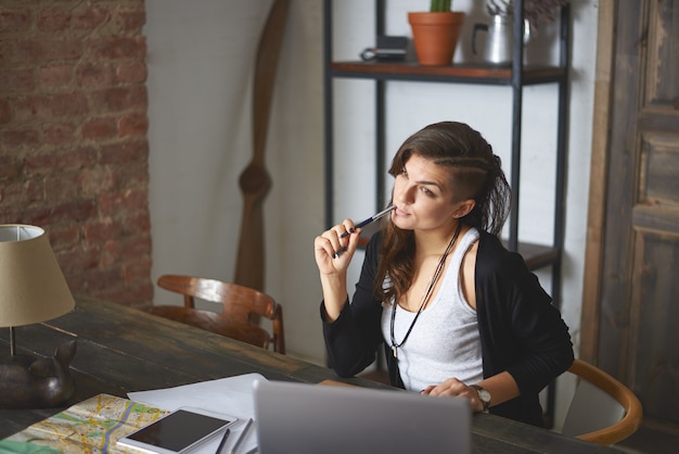 Horizontal indoor shot of beautiful young woman with stylish haircut working in home office, sitting in front of generic laptop computer, having thoughtful look, dreaming about vacations by the sea