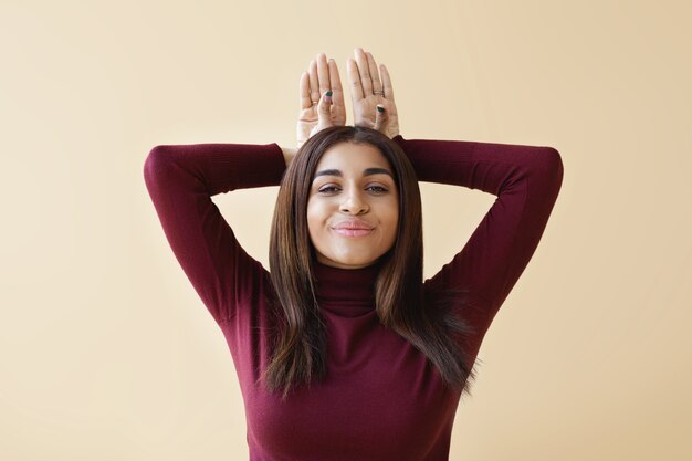 Horizontal isolated shot of attractive positive young mixed race female wearing stylish clothes smiling cheerfully and keeping hands behind her head, imitating bunny while having fun