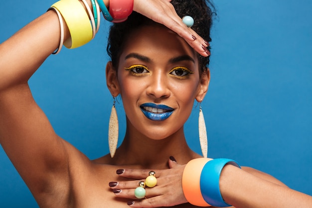 Horizontal lovely mulatto woman with colorful makeup and curly hair in bun showing accessories on her arms isolated, over blue wall