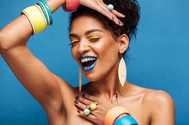 Horizontal lovely mulatto woman with colorful makeup and curly hair in bun smiling and demonstrating accessories on her arms, isolated over blue