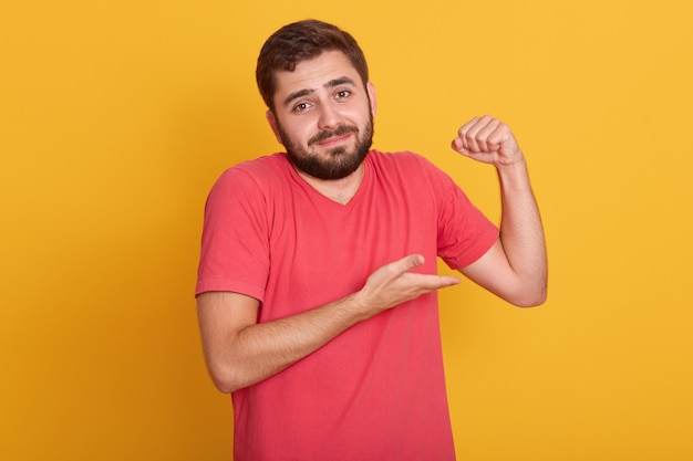 Horizontal male in sleeveless t shirt show his weak biceps muscles, young handsome unshaven man posing isolated on yellow wall, attractive guy with beard.