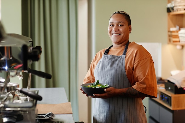 Horizontal medium portrait of cheerful young african american woman with stylish short hair wearing apron standing in cafe kitchen holding bowl with salad smiling at camera