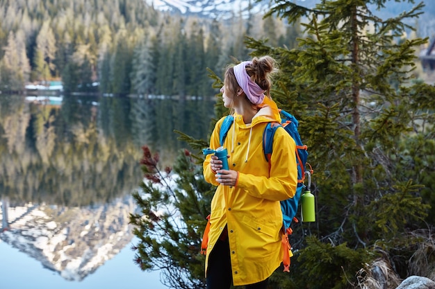 Horizontal outdoor view of calm female tourist admires beautiful mountains, forest and lake, keeps gaze aside