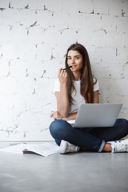 Horizontal photo of cute trendy brunette, sitting on floor with crossed feet in casual clothes, holding laptop, surrounded with notes while biting pen. Prepares for University exams