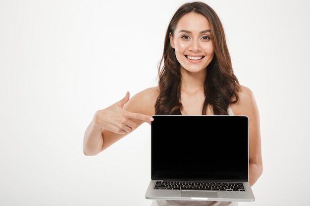 Horizontal photo of happy woman 30s smiling and demonstrating black empty screen of silver laptop on camera with finger, over white wall