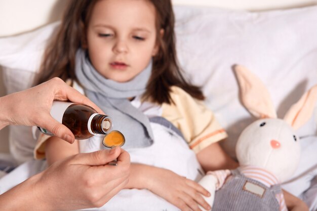 Horizontal photo of unknown hand pouring liquid into spoon from little bottle with syrup, person caring about child lying in bed with flu