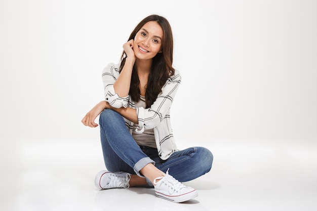 Foto gratuita l'immagine orizzontale della donna castana con capelli castani che si siedono con le gambe ha attraversato sul pavimento e guardare sulla macchina fotografica con il sorriso, isolata sopra la parete bianca