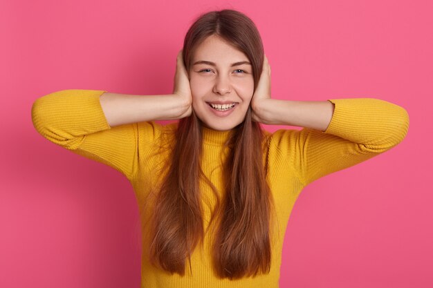 Horizontal picture of cheerful good looking positive young lady having pleasant facial expression, being in good mood, wearing casual clothes, covering ears with hands.