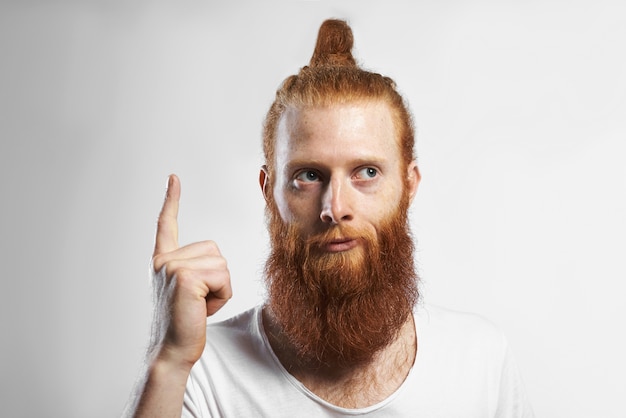 Free photo horizontal picture of emotional funny young unshaven male with trendy hairstyle and stubble having good idea, raising finger up and looking away with pensive mysterious smile. body language