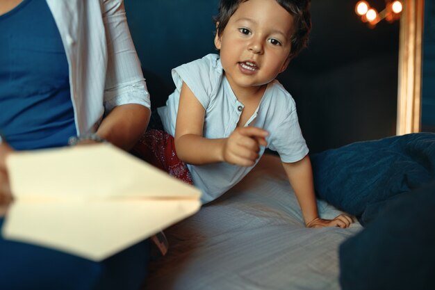 Horizontal Portrait of adorable active little boy playing in bedroom, pointing finger at front