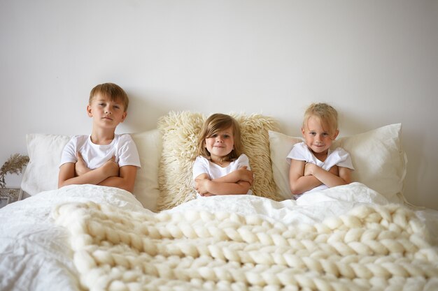 Horizontal portrait of adorable pretty baby girl relaxing in bed between her two elderly brothers. Charming European children siblings crossing arms, refusing to get up early in the morning