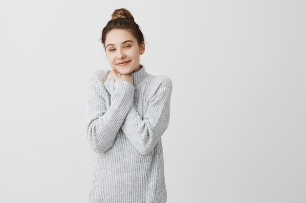 Horizontal portrait of adorable woman tied her hair in topknot smiling being positive. Beautiful female hairdresser having break putting hands on chest in joy. Leisure, person concept