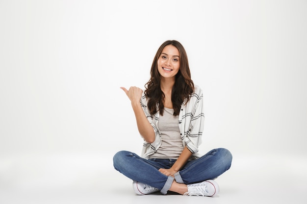 Horizontal portrait of affable woman in casual clothes sitting in lotus pose on the floor and pointing finger aside, isolated over white wall