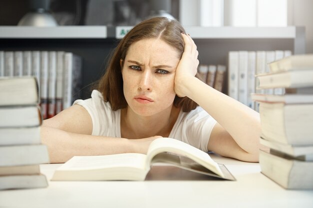 Horizontal portrait of angry, sad and frustrated teenage woman wearing casual clothes and daily make-up, bored with studying a scientific manual at school library, having displeased look and bad mood