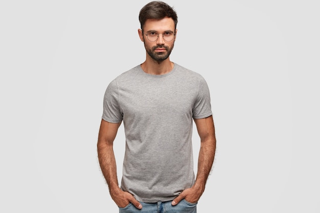 Horizontal portrait of attractive bearded male with serious expression, dressed in casual grey t-shirt, keeps hands in pockets, shows new clothes, isolated over white wall. People, style concept