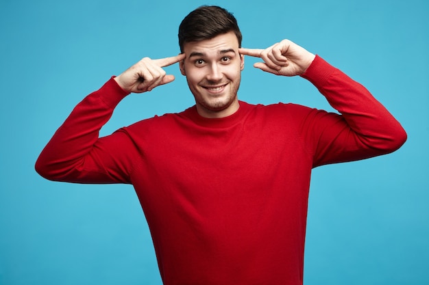 Горизонтальный портрет привлекательного эмоционального молодого темноволосого парня в красном свитере, держащего передние пальцы