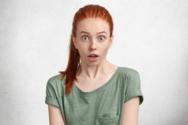 Horizontal portrait of beauitful stunned freckled ginger female in casual t shirt, has bated breath