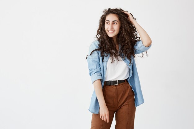 Horizontal portrait of beautiful good looking girl with appealing appearance smiles happily, wears her long wavy hair loose, has good mood after spending weekends with friends, glad and pleased