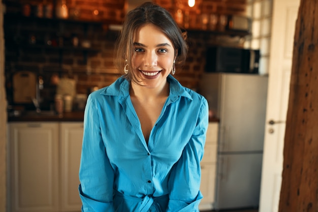 Горизонтальный портрет красивой позитивной молодой европейской женщины в синем платье-рубашке, расслабляющейся дома, глядя вперед с легкой беззаботной улыбкой