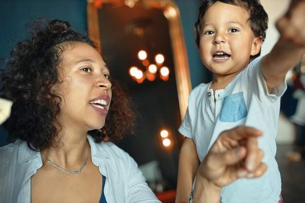 Horizontal Portrait of beautiful young mixed race woman with curly hair having fun indoors together with her excited adorable baby boy