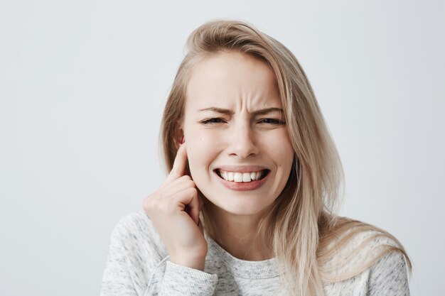 Horizontal portrait of blonde caucasian female dressed casually has headache after noisy party, clenches teeth and holds hand behind her ear. Irritated young female expressing negative emotions.