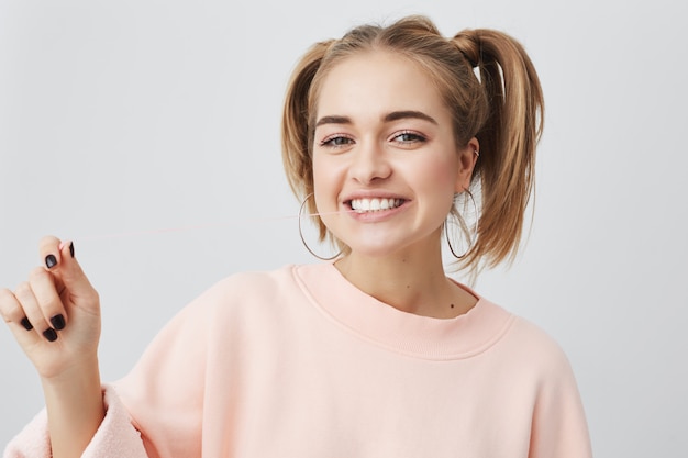 Horizontal portrait of cheerful young woman with appealing smile, demonstrarting white perfect teeth, having two ponytails on her head, wearing pink sweatshirt. Cute girl showing positive emotions