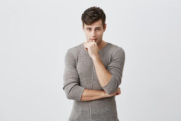 Horizontal portrait of confident serious attractive caucasian man keeping hand under chin, touching lips, wearing casual clothes. Thoughtful male student poses at studio