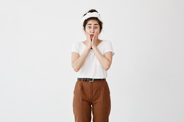 Horizontal portrait of dark-haired stupefied girl keeps mouth opened, with popped eyes, has upset expression, dressed casually, being confused or worried, sees no way out in difficult situation