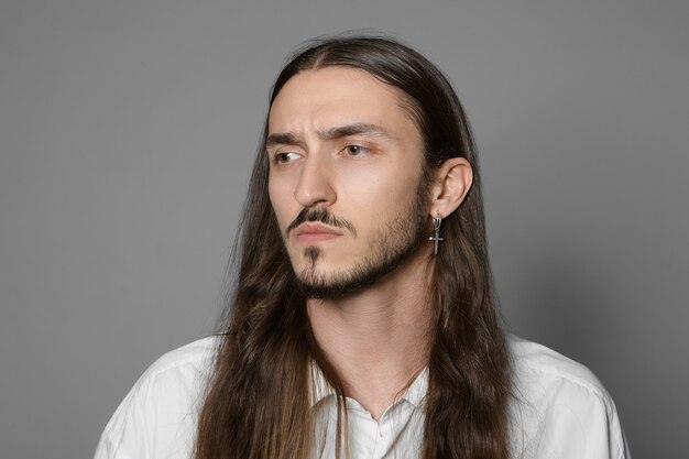 Horizontal portrait of fashionable young unshaven man with earring and long loose hairstyle frowning eyebrows, being worried about work, posing in white shirt