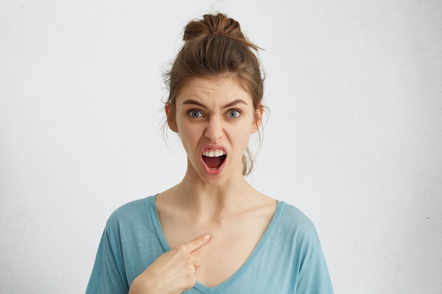 Horizontal portrait of furious young woman with blue eyes having wide opened mouth from anger pointing at herself with finger frowning her eyebrows. Have I done it? People and emotions concept