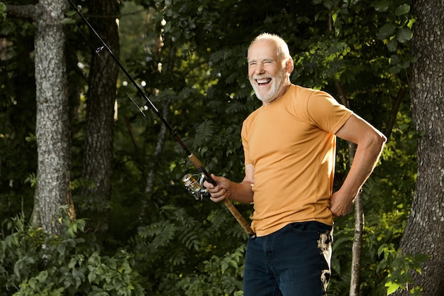 Horizontal portrait of handsome cheerful elderly Caucasian male pensioner in casual clothes laughing happily while standing outdoors with fishing rod, catching fish on river bank in the morning