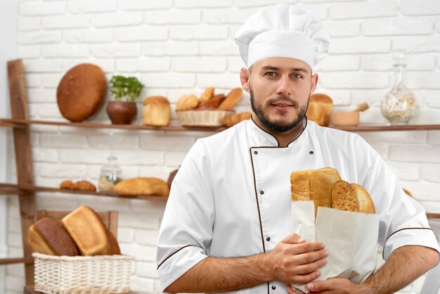 Горизонтальный портрет красивого молодого пекаря, радостно улыбающегося, позирующего в своей пекарне со свежеиспеченным хлебом в бумажном пакете.