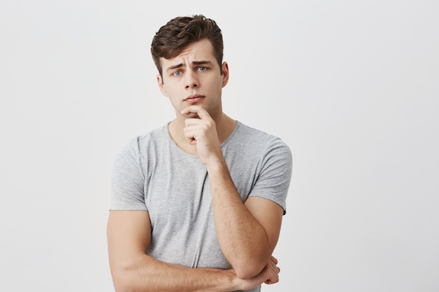 Horizontal portrait of handsome young male dressed casually keeps hand under chin, frowns his face, looks puzzled, listens to someone`s official speech, analyzes information. Peope and lifestyle