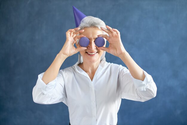 Horizontal portrait of happy gray haired retired Caucasian female in white shirt and conical hat holding two round almond cookies at her eyes