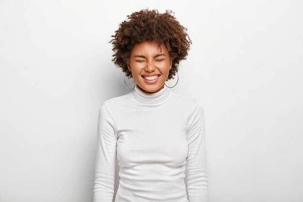 horizontal portrait of joyous afro female laughs loudly, keeps eyes closed, being in good mood, wears turtleneck, isolated on white wall. people, satisfaction, pleasure, ethnicity, emotions