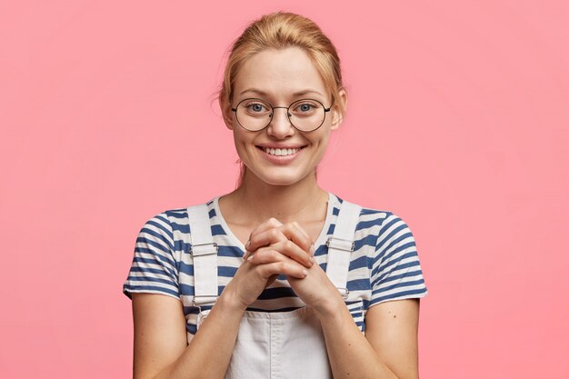 Horizontal portrait of lovely European female wears casual striped t shirt and denim dungarees, keeps hands together