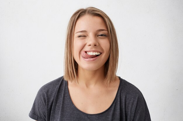 Horizontal portrait of optimistic funny girl with bob hairstyle showing her tongue and blinking eyes