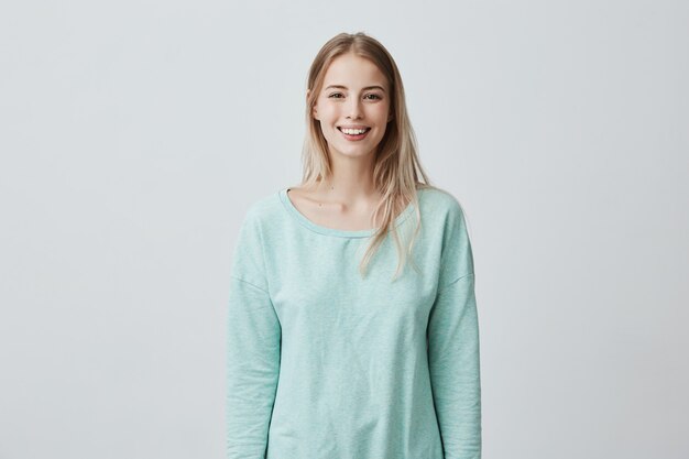 Horizontal portrait of pleasant-looking Caucasian female with long blonde hair, wearing white casual blue sweater, looking happily