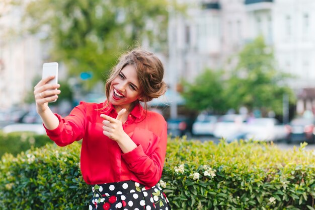 Horizontal portrait of pretty girl standing in park. She wears red blouse and nice hairstyle. She is making a selfie-portrait on phone.