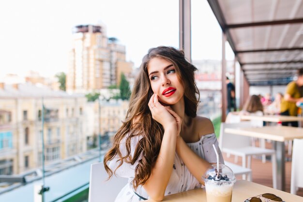 Horizontal portrait of pretty girl with long hair sittting at table on the terrace in cafe. She wears a white dress with bare shoulders and red lipstick . She is looking to the side.